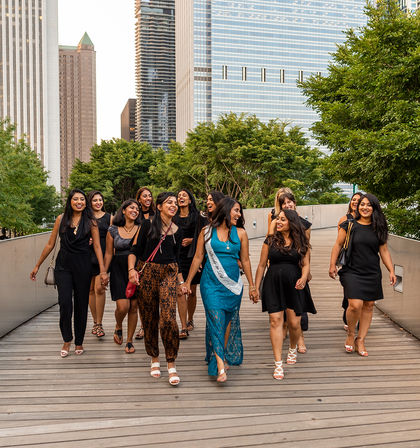 Group of women on a bachelorette outing, bride-to-be wearing a sash, walking and laughing on an urban wooden promenade with downtown skyscrapers and trees