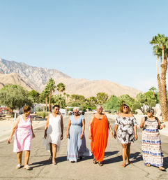 Six women in colorful summer dresses walking down a sunlit desert street with palm trees and rocky mountains in the background, relaxed vacation vibe