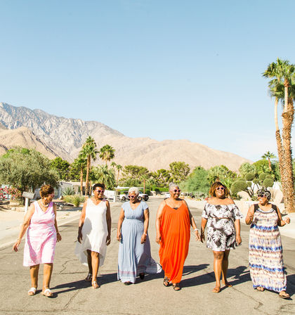 Six women in colorful summer dresses walking down a sunlit desert street with palm trees and rocky mountains in the background, relaxed vacation vibe