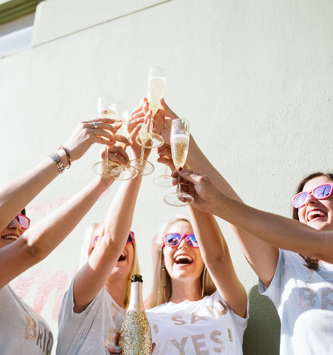 Group of women in matching tees and pink sunglasses cheering with champagne flutes and a glitter-covered bottle against a sunlit wall — playful bachelorette toast