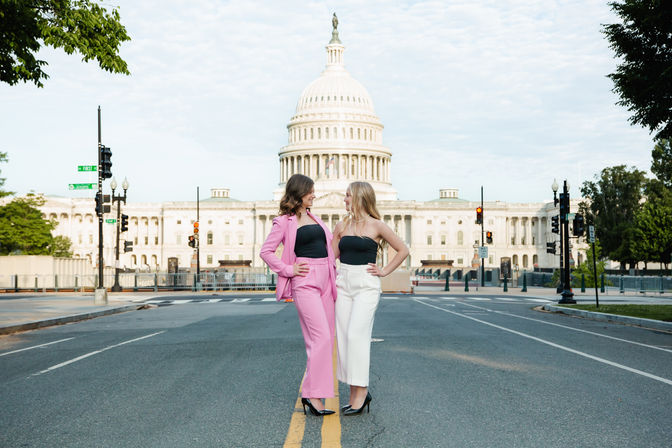 Two women pose on an empty street in Washington, D.C., in front of the U.S. Capitol dome—one in a pink suit and the other in white pants with black tops, smiling and facing each other.