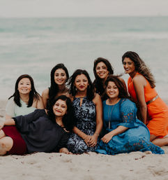 Seven women smiling and posing on a sandy beach with the ocean behind them, wearing colorful summer dresses.