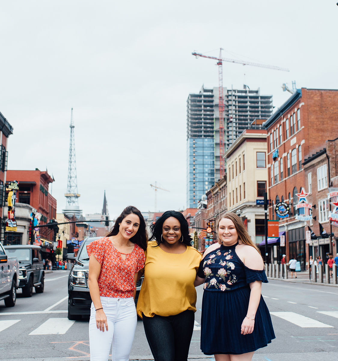 Three women smiling and walking arm‑in‑arm down a lively downtown city street lined with brick storefronts, neon signs, and a high‑rise under construction in the skyline.