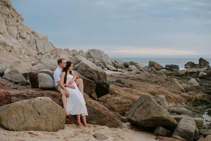 Romantic couple barefoot on a rocky coastline — woman in a white dress and man in a light shirt embrace on coastal boulders with the ocean and cloudy sky in the background.