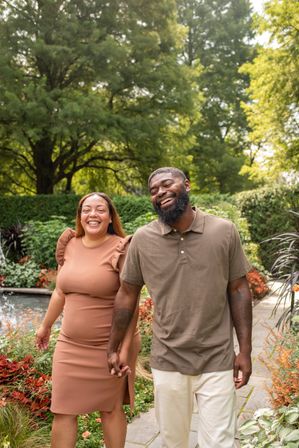 Smiling couple strolling hand-in-hand along a lush botanical garden path surrounded by trees, colorful flowers, and a small pond.