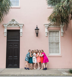 Six friends in colorful dresses posing on a sidewalk in front of a pastel pink historic facade with white trim, black lantern and palm trees.