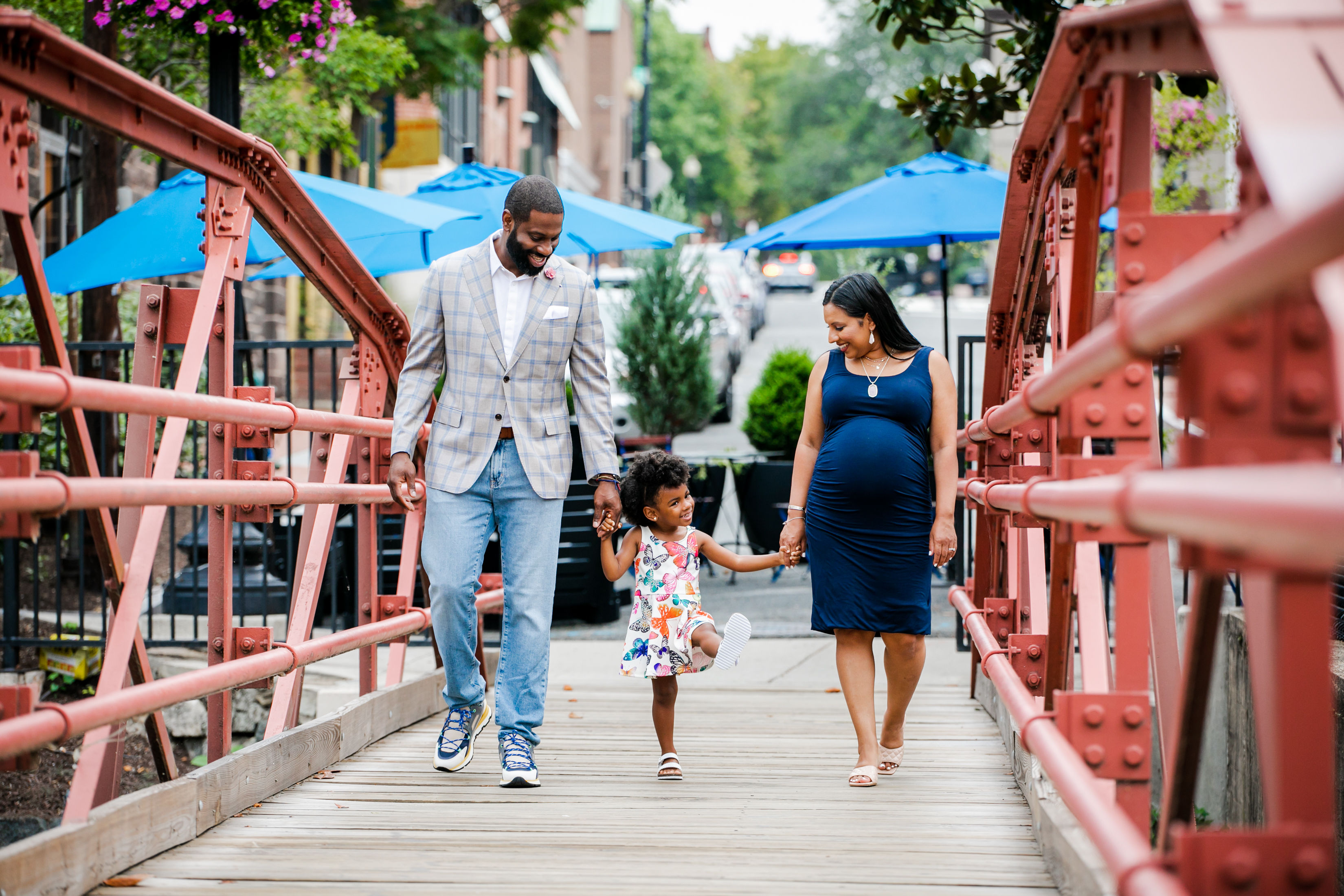 Pregnant woman, man, and young daughter holding hands and joyfully walking across a red metal pedestrian bridge in a downtown streetscape with blue patio umbrellas.