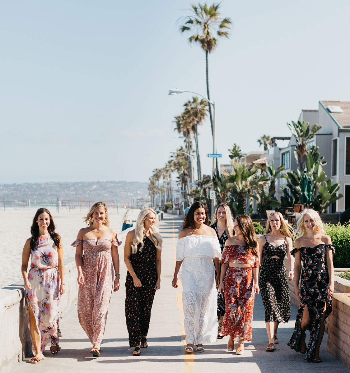 Eight women in colorful summer dresses walking along a sunny California beach boardwalk lined with palm trees and ocean views.