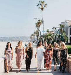 Eight women in colorful summer dresses walking along a sunny California beach boardwalk lined with palm trees and ocean views.