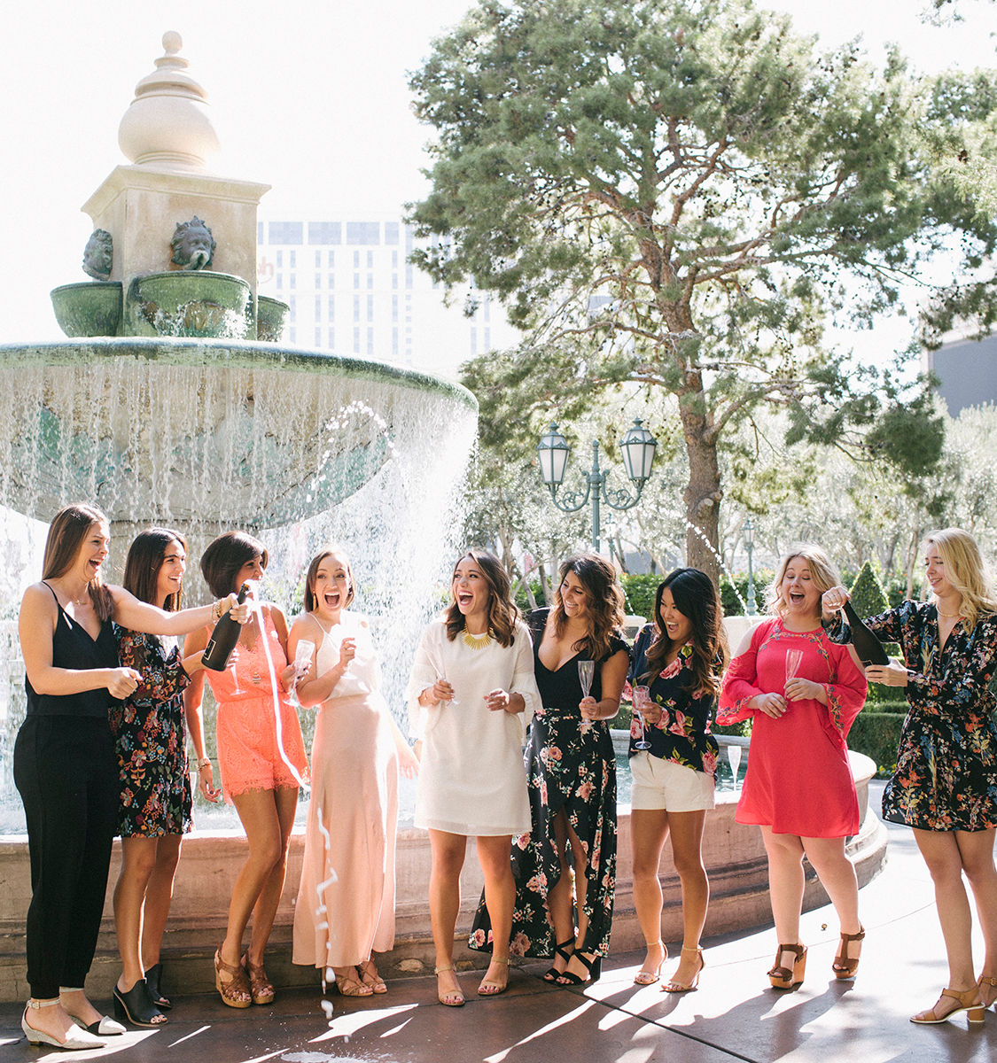 Group of women in colorful summer dresses laughing and popping champagne beside a large decorative fountain in a sunny urban courtyard — friends celebrating outdoors with raised glasses and festive energy.