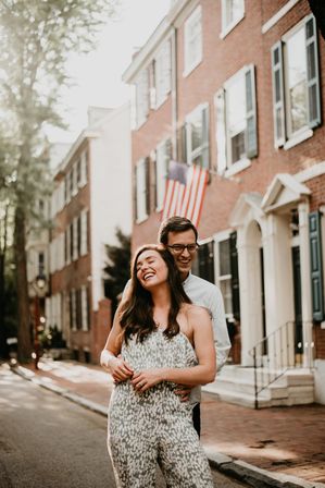 Happy couple embracing on a sunlit historic brick rowhouse street with shuttered windows, stoops and an American flag in the background