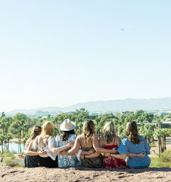 Six friends sitting arm-in-arm on a rocky overlook, overlooking a palm-tree-lined desert park with a lake and distant mountains under a clear blue sky — sunny outdoor reunion vibe.
