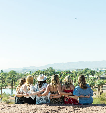 Six friends sitting arm-in-arm on a rocky overlook, overlooking a palm-tree-lined desert park with a lake and distant mountains under a clear blue sky — sunny outdoor reunion vibe.