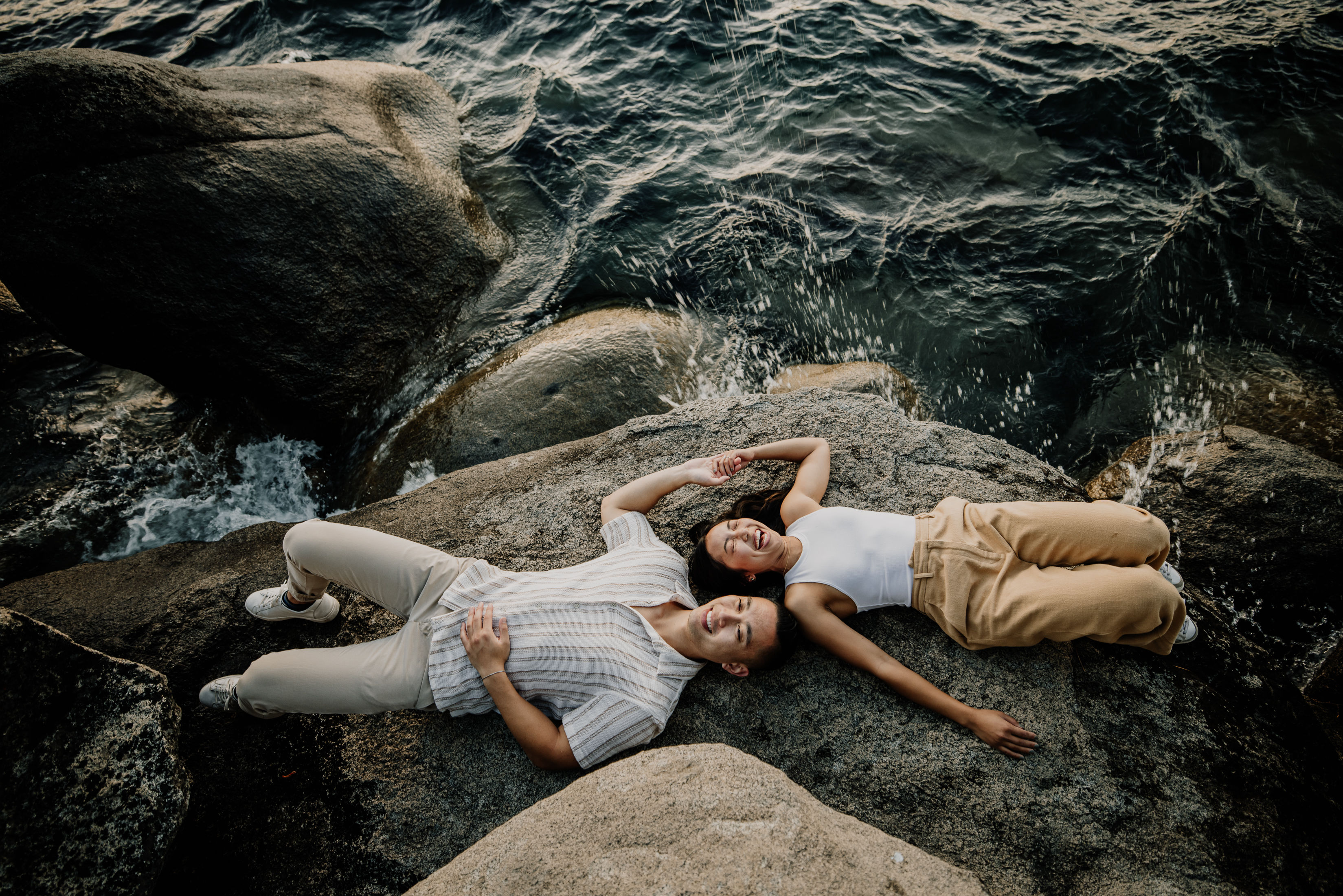 Overhead view of two people lying and smiling on a rocky shoreline, holding hands as ocean waves splash against the rocks.