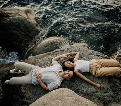 Overhead view of two people lying and smiling on a rocky shoreline, holding hands as ocean waves splash against the rocks.
