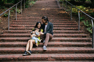 Young family — mother, father and toddler — cuddling on wide brick park steps with metal handrails and surrounding ferns, smiling.
