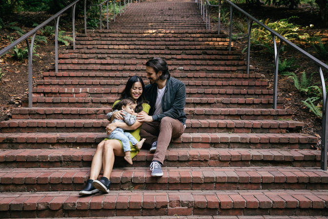 Young family — mother, father and toddler — cuddling on wide brick park steps with metal handrails and surrounding ferns, smiling.