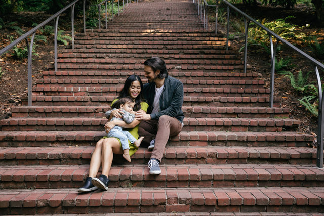 Young family — mother, father and toddler — cuddling on wide brick park steps with metal handrails and surrounding ferns, smiling.