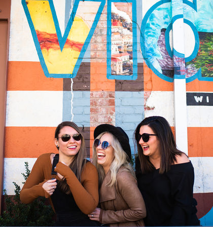 Three laughing women in sunglasses and fall jackets pose in front of a colorful street-art mural with large painted letters and orange-and-white stripes