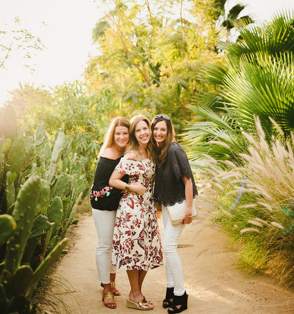 Three smiling women posing on a sunny botanical garden path flanked by cacti, palm fronds, and ornamental grasses, wearing summer dresses and sandals.