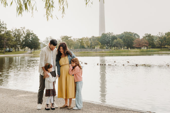 Smiling family of four—pregnant mother, father and two daughters—standing by a lake in an urban park with a tall obelisk tower reflected in the water, trees lining the shore