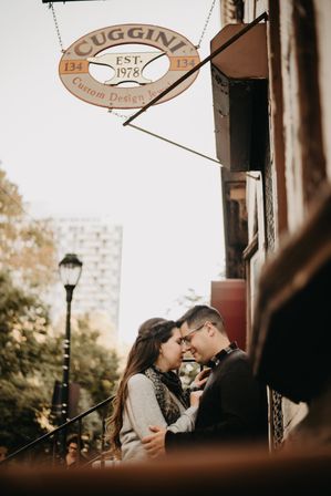Romantic couple forehead-to-forehead on a city stoop beneath a hanging vintage shop sign, soft-focus lamppost, trees, and urban storefront in autumn tones.