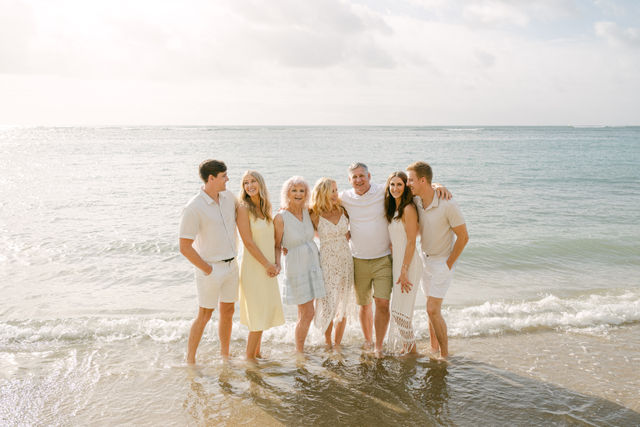 Seven-person multigenerational family in light summer outfits standing barefoot in shallow ocean water, smiling and embracing on a sunny beach with calm waves and a pale blue sky.
