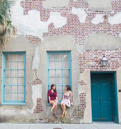 Two women in summer dresses sit and chat on a window ledge of a weathered brick-and-plaster facade with teal-framed windows and door on an urban sidewalk.