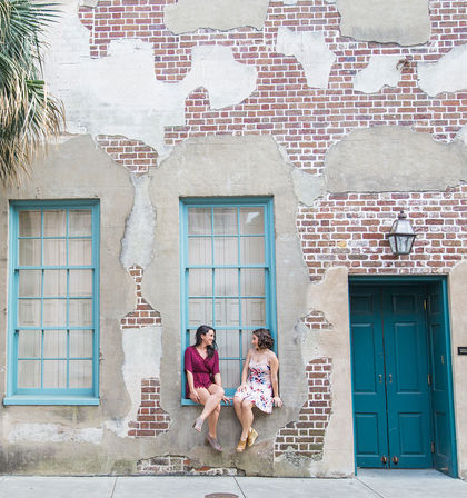 Two women in summer dresses sit and chat on a window ledge of a weathered brick-and-plaster facade with teal-framed windows and door on an urban sidewalk.