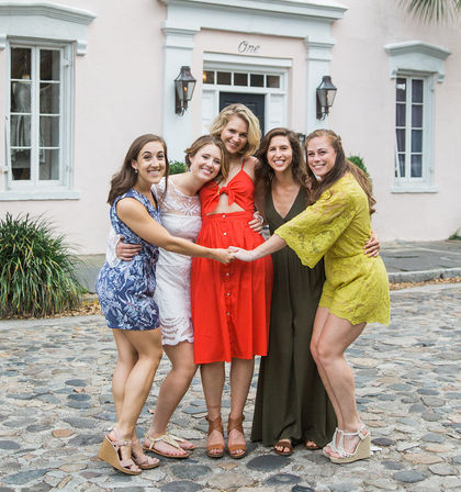 Five friends in colorful summer dresses laughing and hugging on a cobblestone street in front of a charming pastel pink house with white trim and lanterns