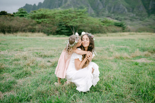 Mother and daughter wearing floral crowns embrace in a lush green meadow with jagged mountains in the background; little girl in a pink dress kisses woman in a white dress.