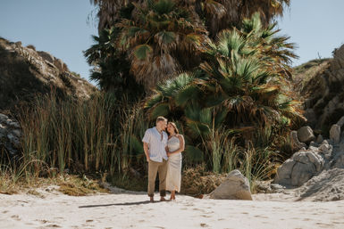 Romantic couple on a sunlit sandy beach cove framed by lush palm trees, tall grasses and rocky cliffs