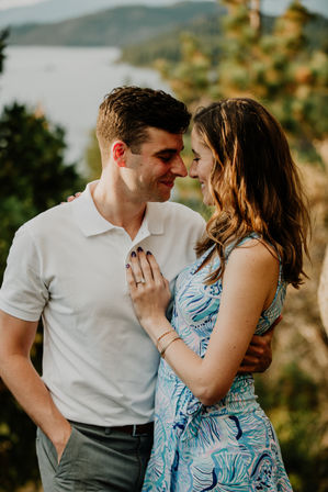 Sunlit lakeside engagement-style portrait of a smiling couple in a warm embrace, woman in a blue patterned dress showing an engagement ring with forested hills in the background.