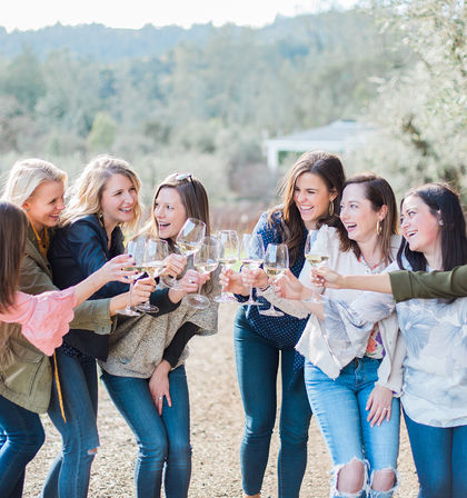 Group of friends laughing and clinking white-wine glasses during an outdoor wine tasting in a sunlit vineyard setting