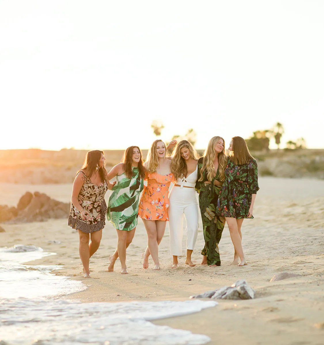 Six women walking barefoot along a sandy beach at golden-hour sunset, laughing arm-in-arm with gentle waves and distant palm trees on the shoreline