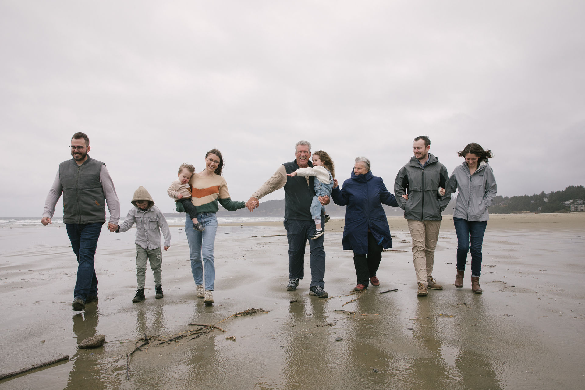 Multigenerational family walking hand-in-hand along a wet sandy beach on an overcast day with waves and a tree-lined coastline in the distance.