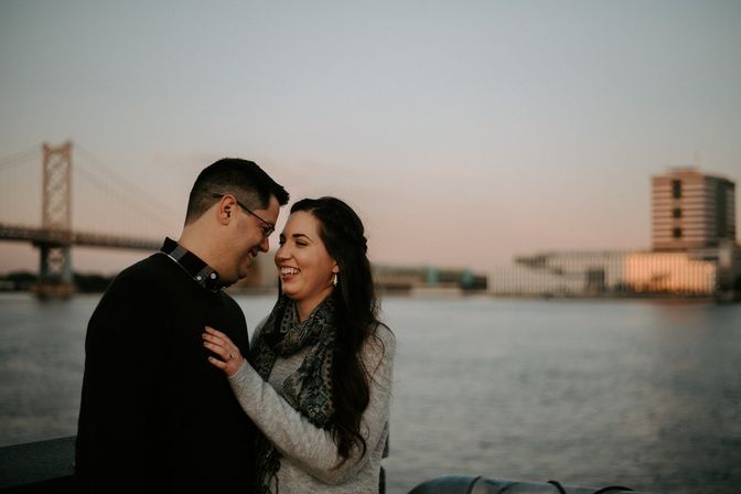 Laughing couple embracing on a riverfront promenade at sunset with a suspension bridge and modern waterfront buildings in the background.
