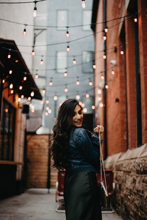 Smiling woman in a denim jacket turns back in a cozy urban alley lit by hanging string lights and warm brick walls, holding a chain-strap handbag.