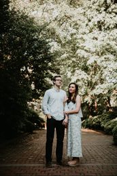 Couple linked arms standing on a sunlit brick pathway through a leafy, tree-lined park — woman in a patterned summer dress and wedges, man in glasses and a casual button-up shirt.