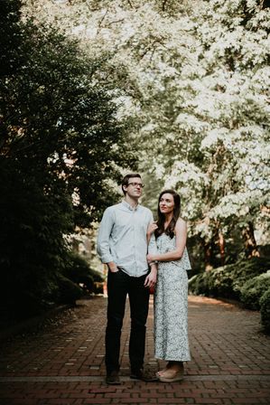 Couple linked arms standing on a sunlit brick pathway through a leafy, tree-lined park — woman in a patterned summer dress and wedges, man in glasses and a casual button-up shirt.