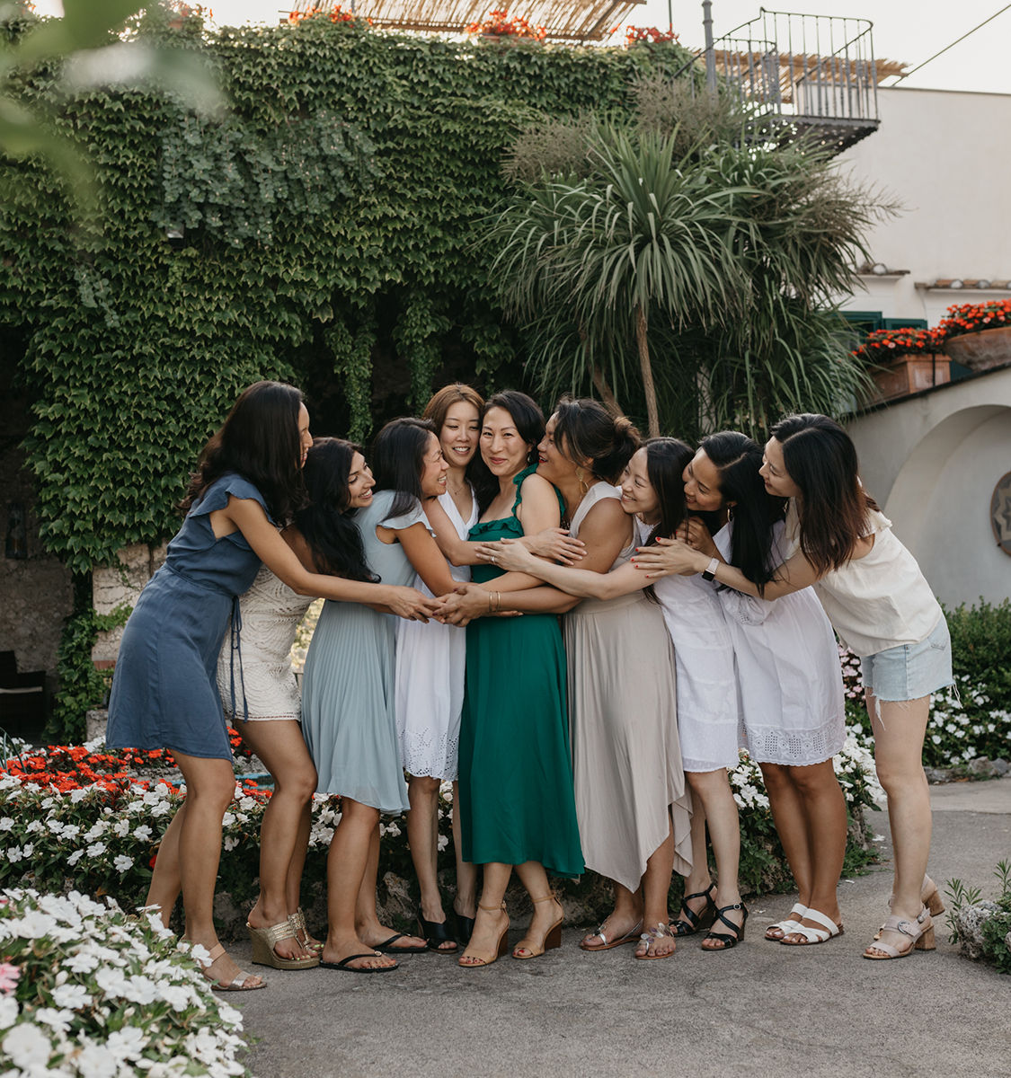 Group of women in summer dresses sharing a joyful group hug in a sunlit garden courtyard with an ivy-covered wall, potted palms and colorful flower beds.