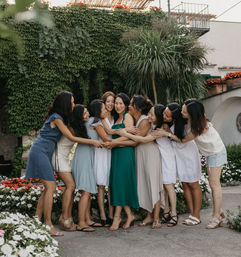 Group of women in summer dresses sharing a joyful group hug in a sunlit garden courtyard with an ivy-covered wall, potted palms and colorful flower beds.