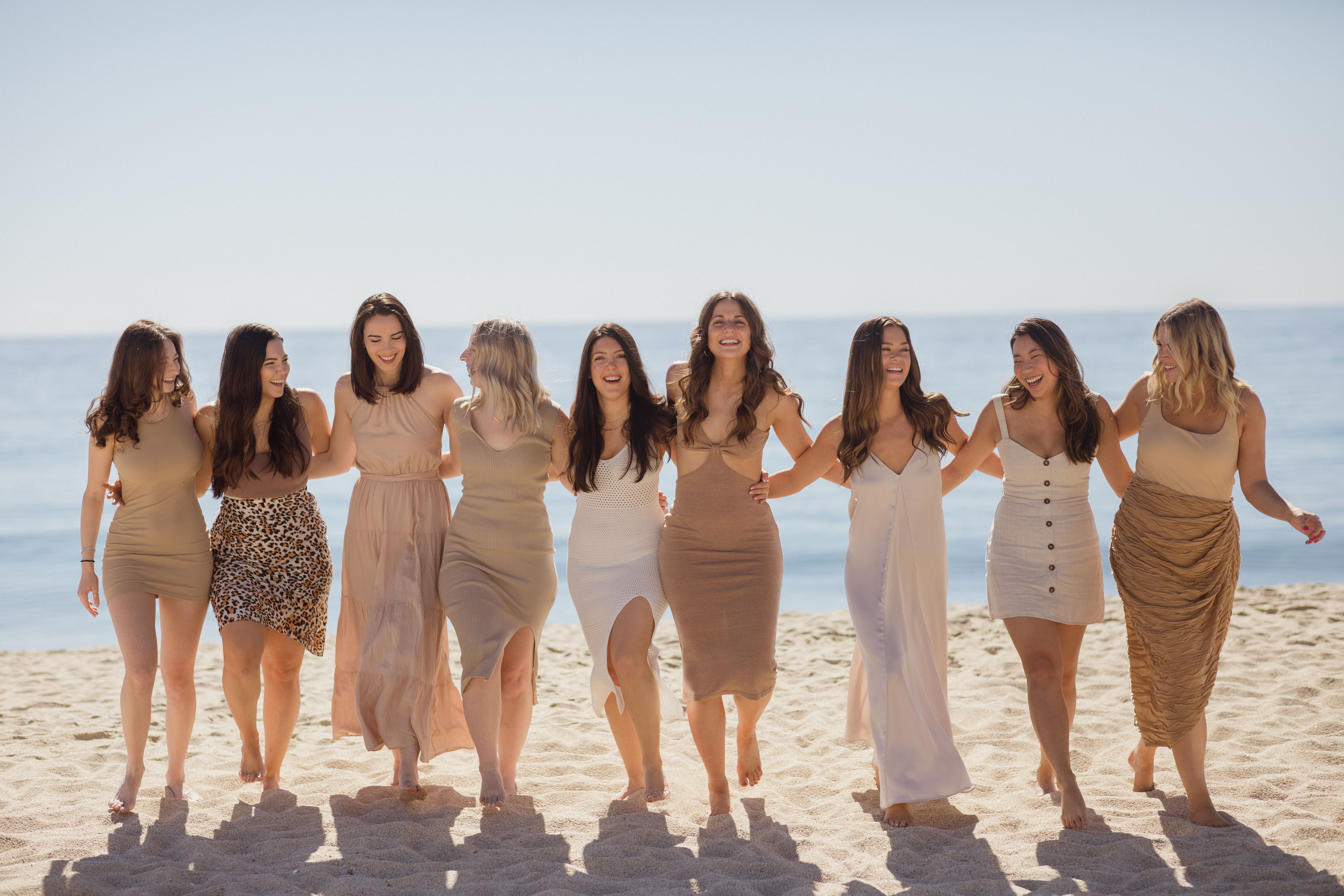 Nine women in neutral-toned summer dresses walking barefoot arm‑in‑arm on a sunny sandy beach with the calm blue ocean in the background