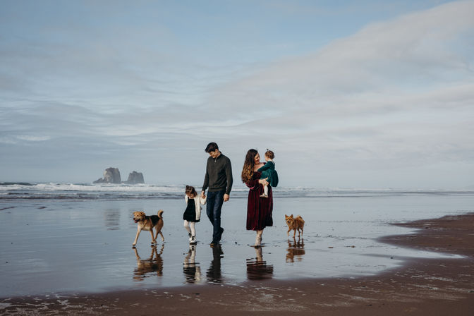 Coastal family photo: two parents and two young children with two dogs walking along a reflective wet sandy beach toward the ocean, distant rock formations and an expansive cloudy sky.