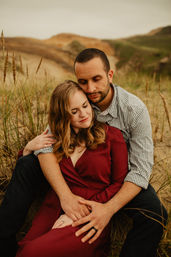Couple cuddling on coastal sand dunes amid tall beach grass — woman in a burgundy dress and man in a checkered shirt in a warm embrace