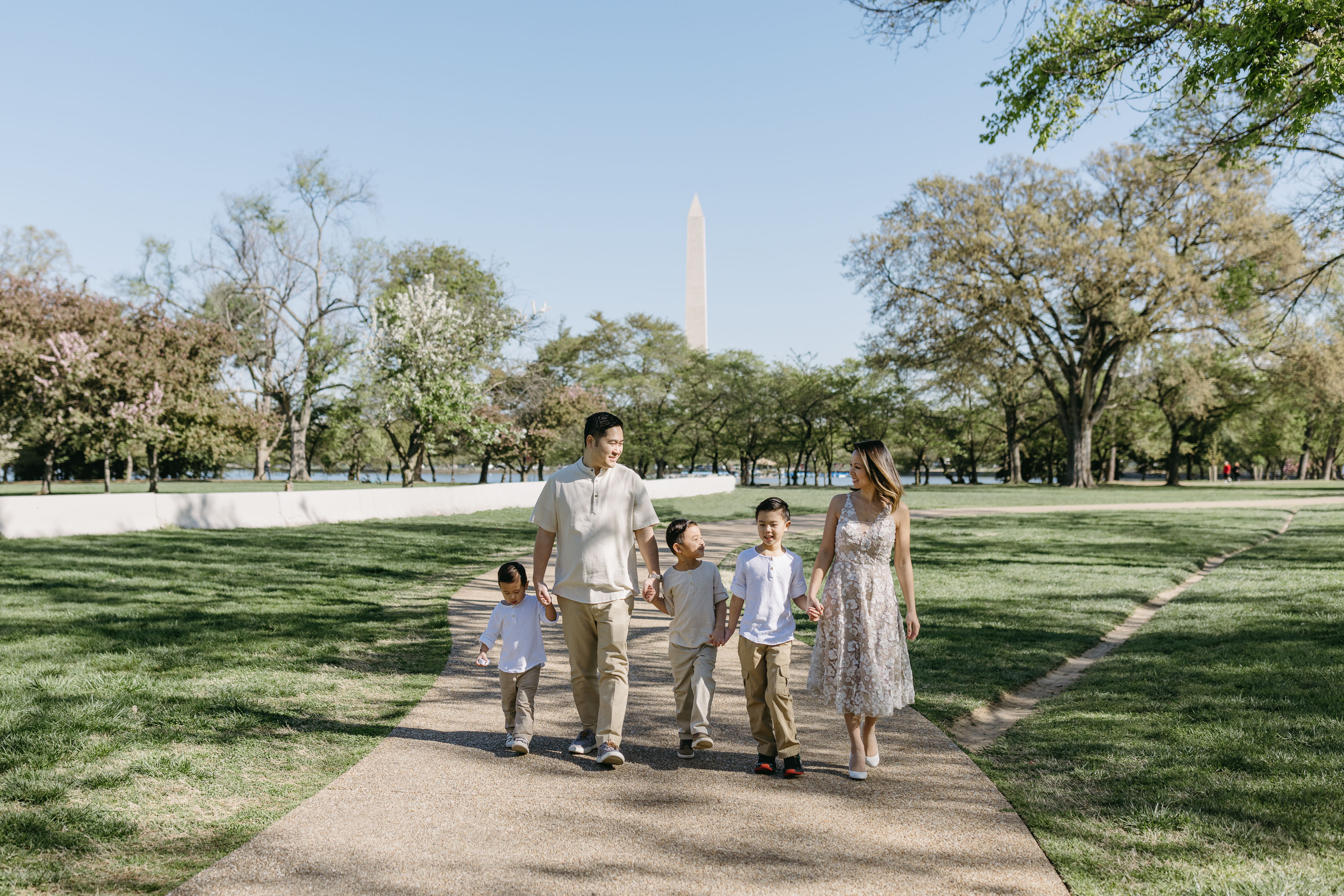 Family of five strolling hand-in-hand on a sunny park path with the Washington Monument in the background, surrounded by spring trees and green lawn.