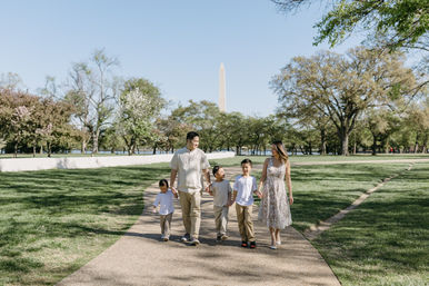 Family of five strolling hand-in-hand on a sunny park path with the Washington Monument in the background, surrounded by spring trees and green lawn.