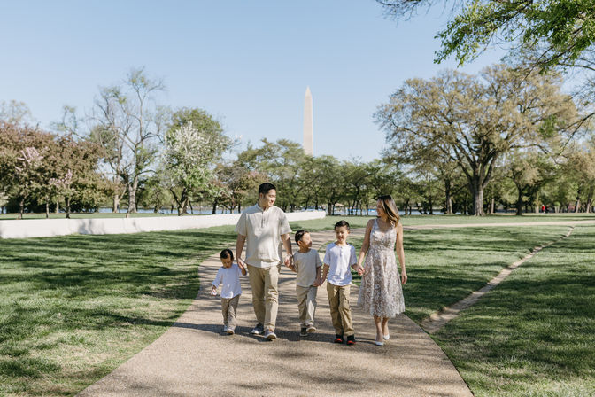 Family of five strolling hand-in-hand on a sunny park path with the Washington Monument in the background, surrounded by spring trees and green lawn.