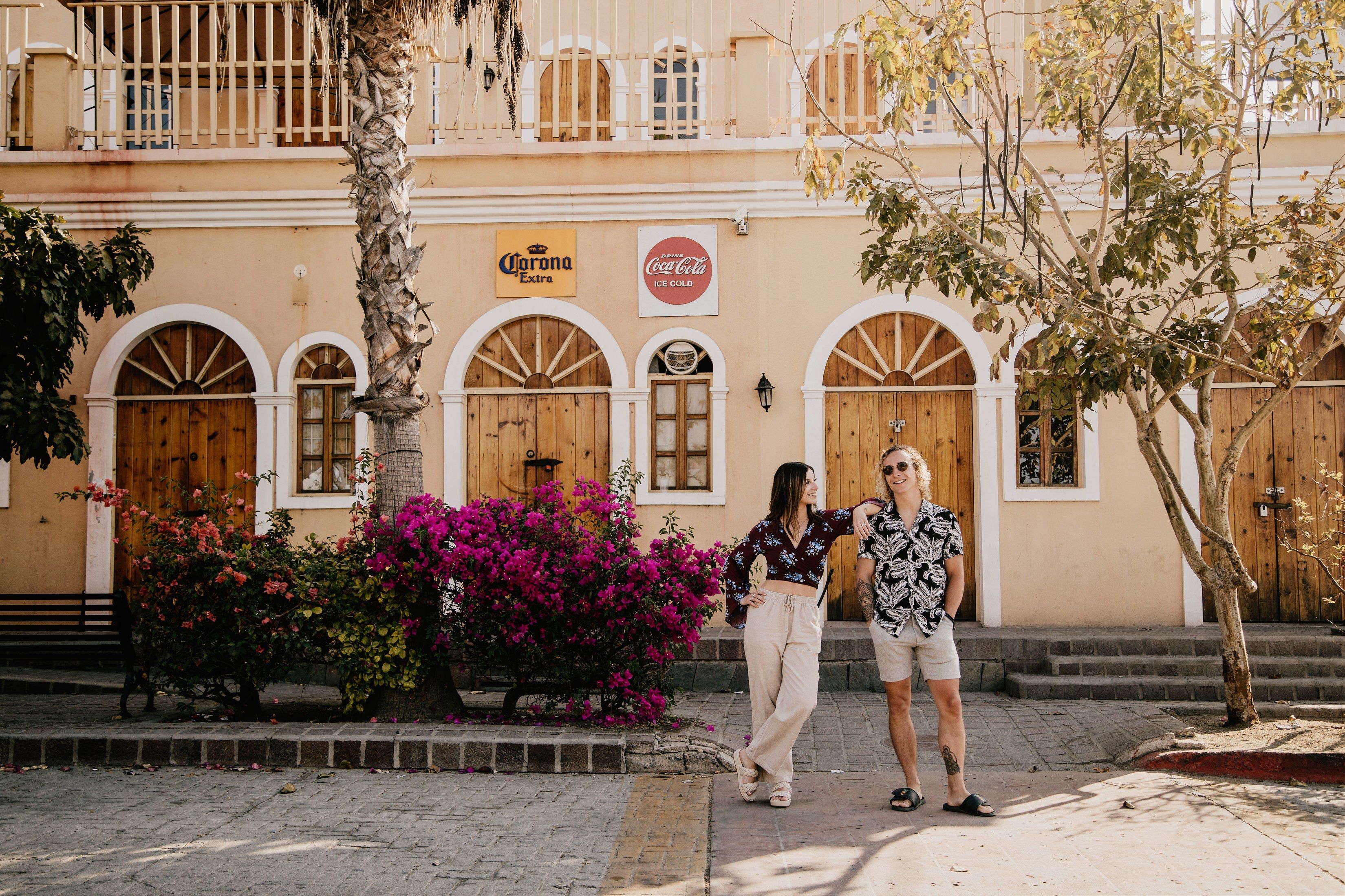 Two friends laughing in a sunlit seaside town plaza in front of a pastel building with arched wooden doors, bougainvillea, and a palm tree