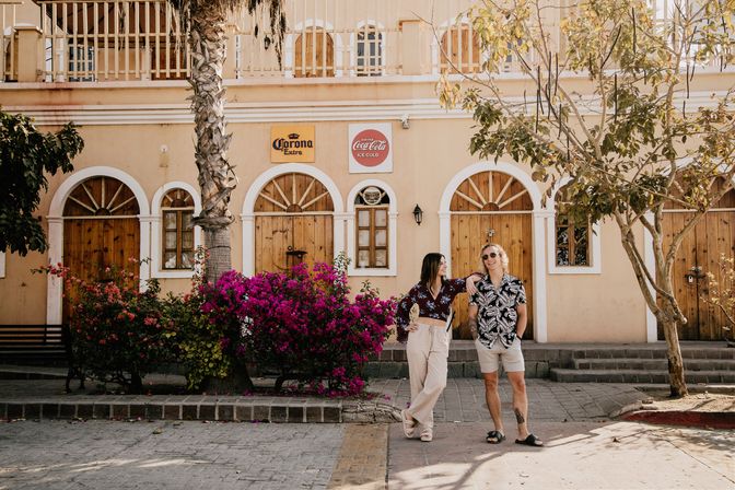 Two friends laughing in a sunlit seaside town plaza in front of a pastel building with arched wooden doors, bougainvillea, and a palm tree